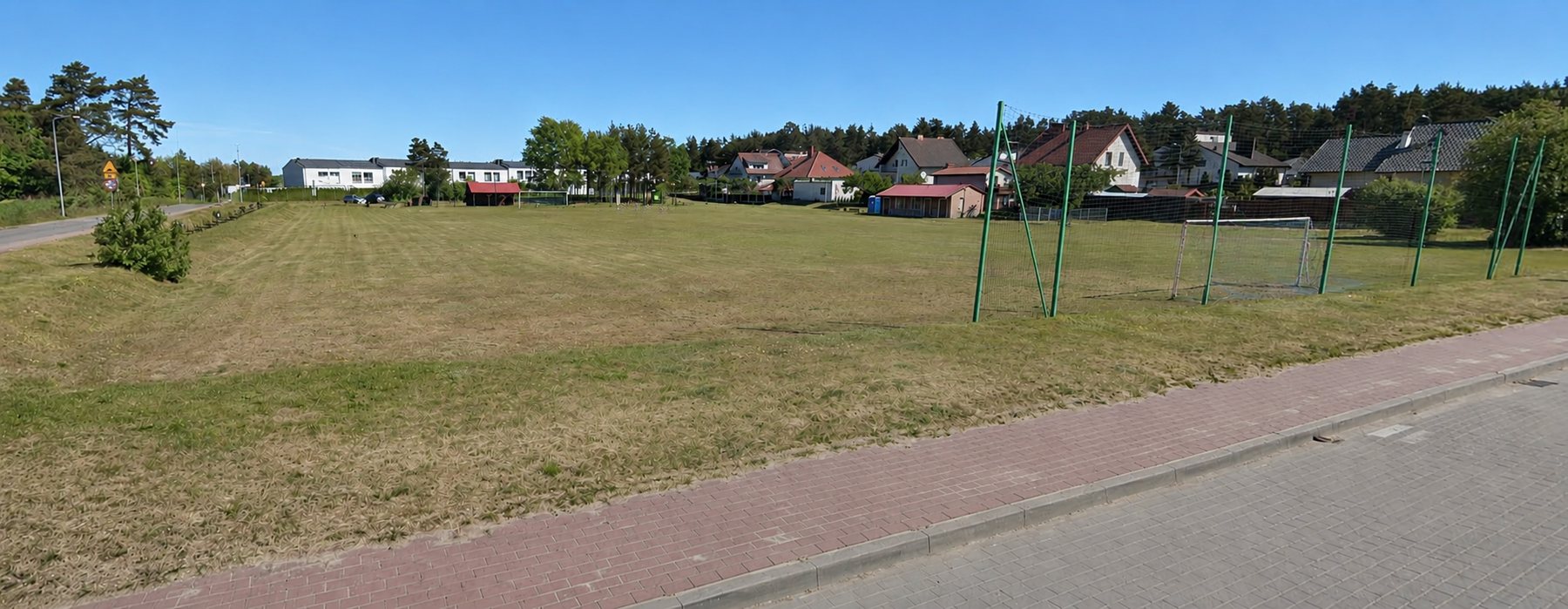 Boisko na osiedlu Trzesieka w Szczecinku — rozległe trawiaste boisko z bramkami i zielonymi piłkochwytami, otoczone domami jednorodzinnymi i lasem
